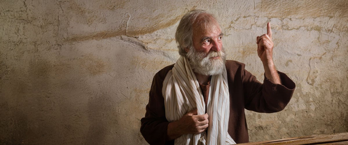 Bearded older man in biblical-style robe and scarf pointing upward against a rough stone wall, portraying a prophet