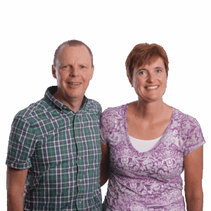 Head-and-torso portrait of Chris & Carole Staples standing side by side, smiling against a plain mauve background