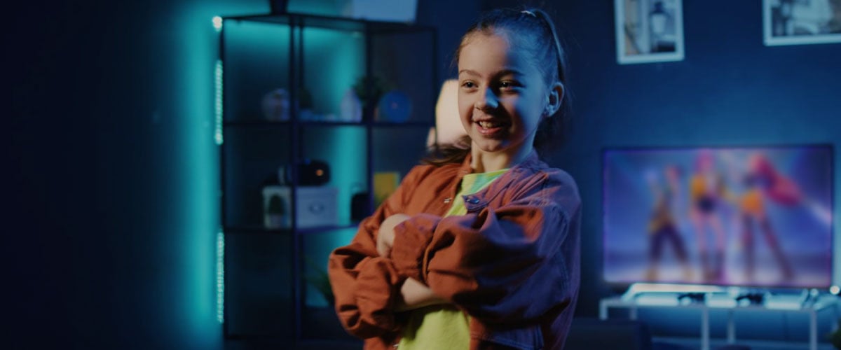 Smiling young girl with arms crossed in a dimly lit room, TV screen showing colourful performers in the background