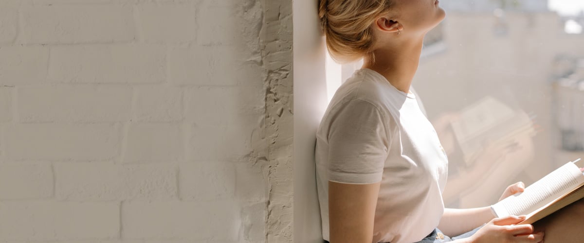 Woman seated by a window, leaning against a wall and reading a book in bright natural light