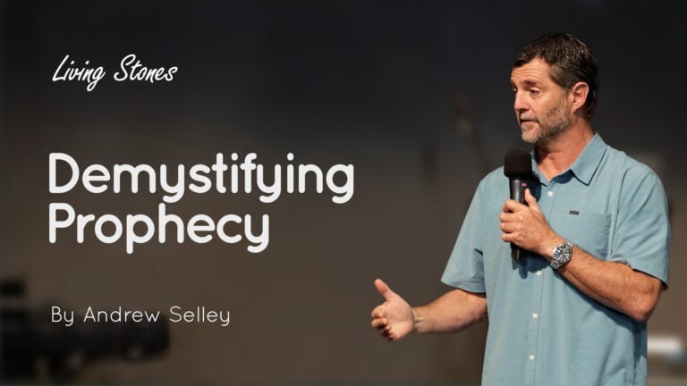 Man holding a microphone speaking on stage beside large title text 'Demystifying Prophecy' and 'By Andrew Selley'