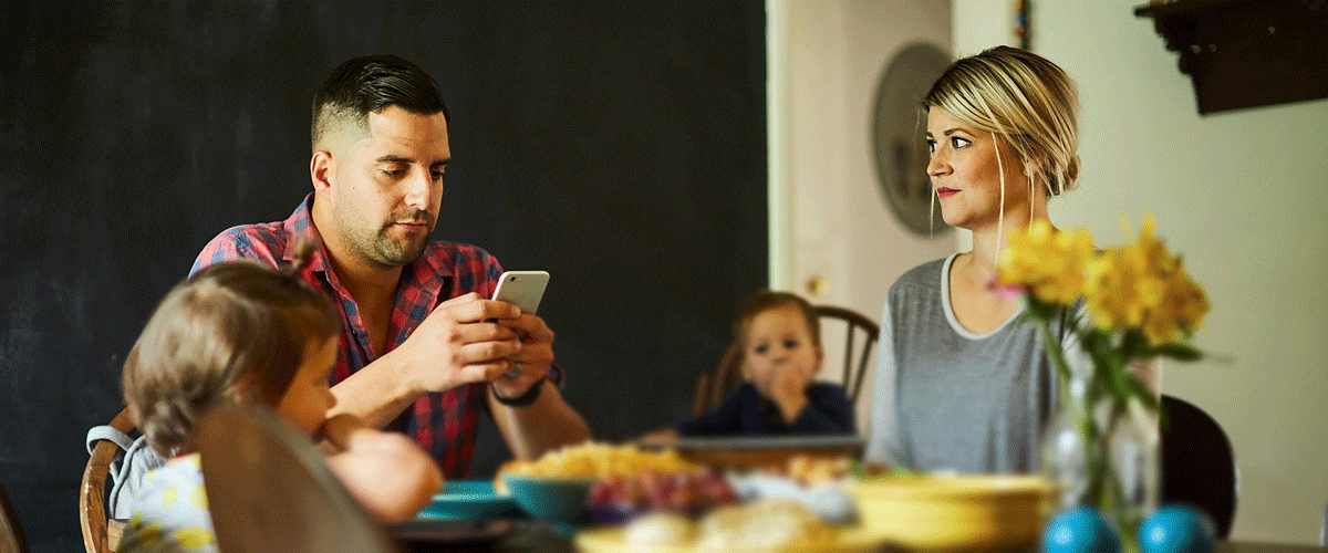 Family at a dining table with father using a smartphone, mother looking concerned and two young children