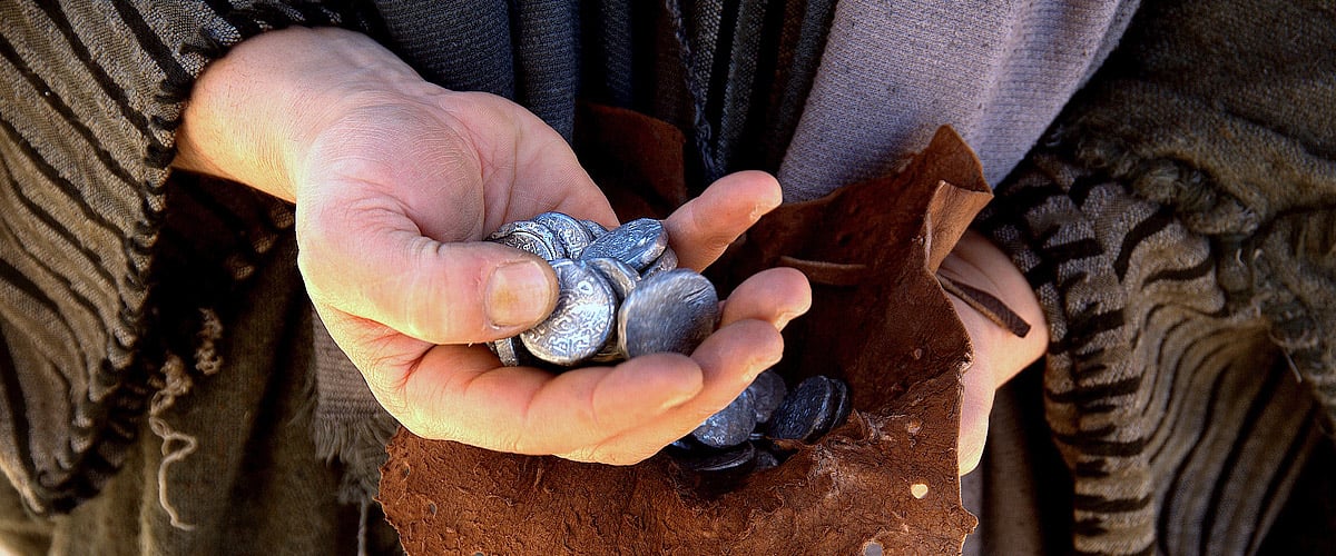 Close-up of hands holding silver coins spilling from a leather pouch, person wearing a dark cloak