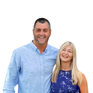 Andrew & Emma Selley standing close and smiling; man in light blue shirt, woman in blue floral dress
