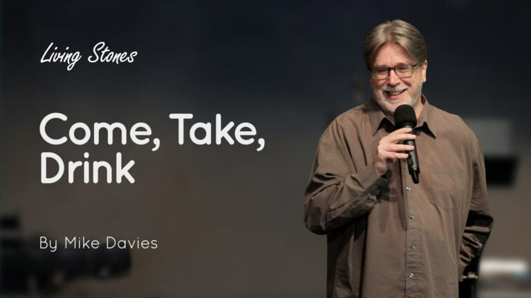 Mike Davies on stage holding a microphone, smiling, with the text 'Come, Take, Drink' displayed beside him