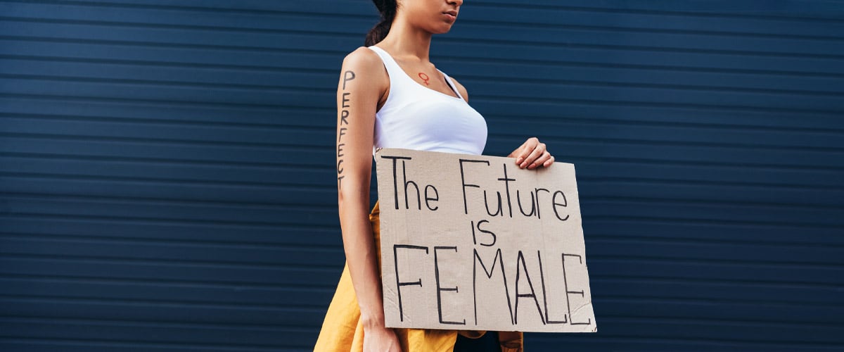 Woman shown from shoulders down holding a cardboard sign reading "The Future is FEMALE" against a blue wall