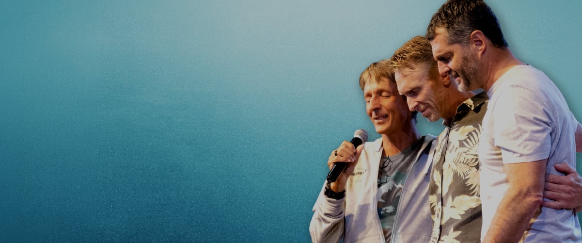 Three men standing close with arms around each other, one holding a microphone against a blue background