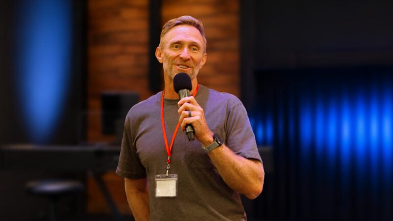 Brad Verreynne speaking into a handheld microphone, wearing a lanyard and casual t-shirt