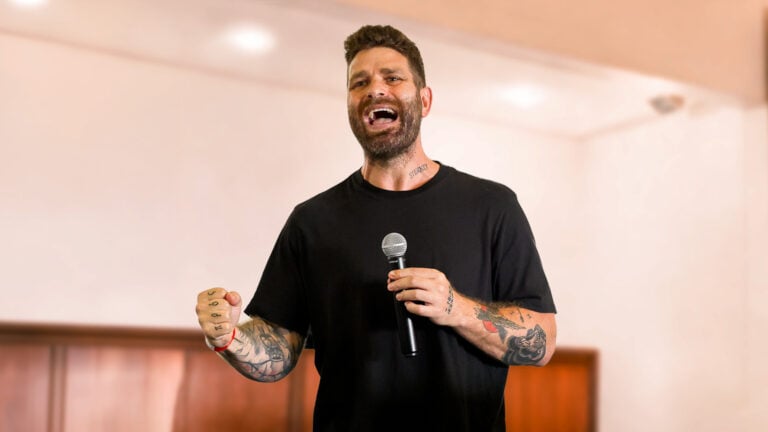 Luke Watson, tattooed man holding a microphone mid-speech, wearing a black T‑shirt