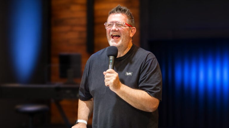 Martin Stockdale holding a microphone on stage, smiling and laughing, wearing glasses and a dark T‑shirt