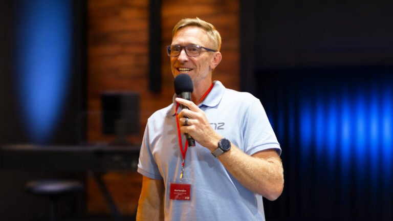 Speaker Brad Verreynne smiling and holding a microphone on stage in a blue polo shirt