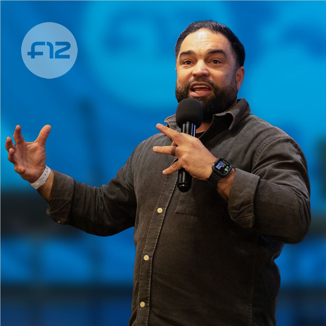 Speaker KevinSamuel holding a microphone and gesturing with both hands against a blue background