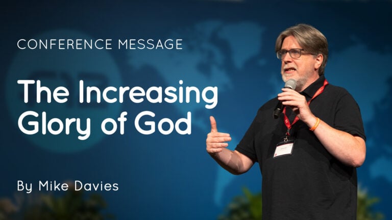 Mike Davies speaking into a microphone on stage beside a slide reading 'The Increasing Glory of God'