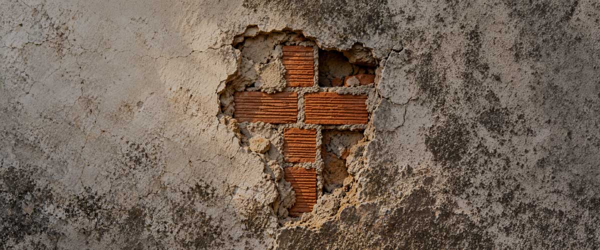 A cross-shaped gap in a weathered, cracked wall reveals red bricks beneath the plaster
