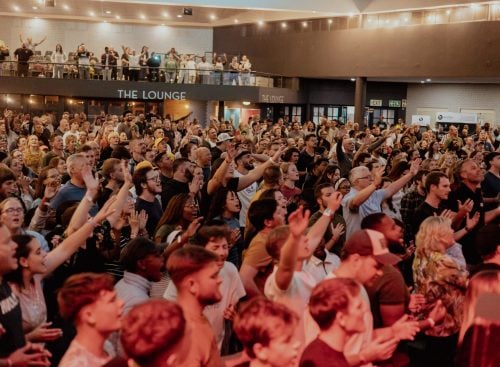 Large indoor crowd with many people standing and raising hands in a hall