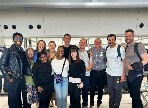 Twelve team members standing side by side in an airport terminal, smiling at the camera