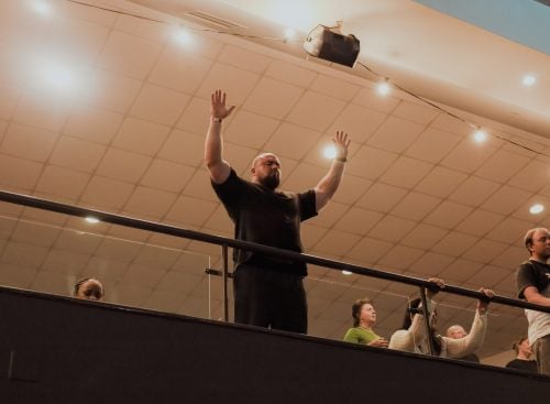 Man standing on an indoor balcony with raised hands, several other people nearby watching