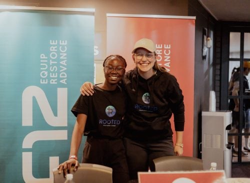 Two people smiling at an event registration table wearing ROOTED shirts with Equip Restore Advance banners behind them