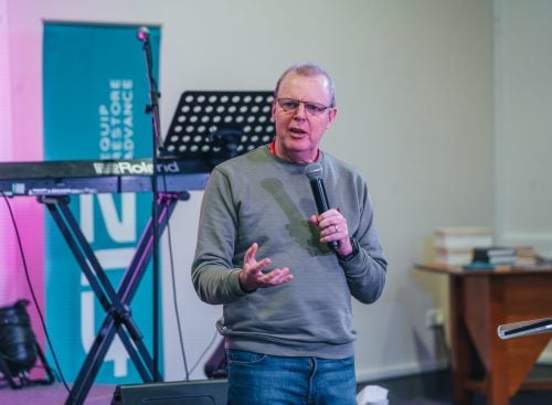 Middle-aged man holding a microphone speaking beside a keyboard and music stand at a leadership event