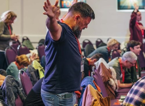 Man standing with arms raised in worship among seated attendees at a 2024 School of Leadership meeting