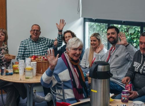 Group of adults sitting around tables waving at the camera during a School of Leadership session