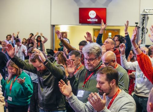 Group of School of Leadership attendees standing with eyes closed and hands raised in a conference room