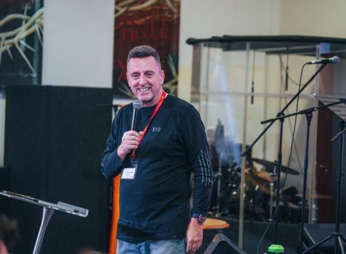 Male speaker smiling and holding a microphone at a lectern during the 2024 School of Leadership event