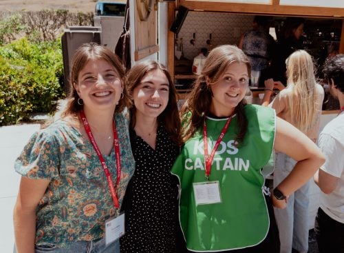 Three smiling young women standing together, one wearing a green 'KIDS CAPTAIN' bib and red conference lanyards