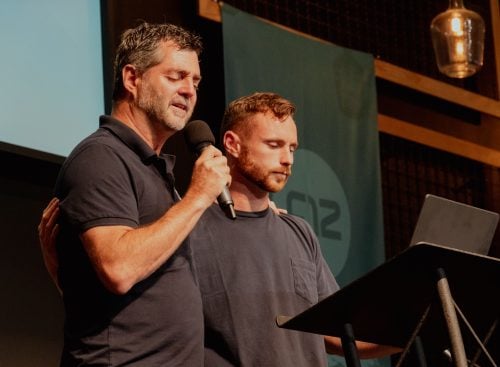 Two men at a lectern during 2025-CONFERENCE-IN-THE-USA-MINISTRY-35; one holds a microphone and has his arm around the other