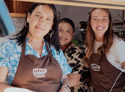 Three smiling volunteers wearing 'Holy Grounds' aprons at a serving window during the 2025 conference in the USA