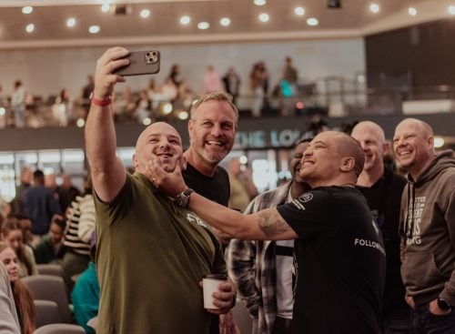 Group of smiling men taking a selfie in a crowded auditorium, one holding a takeaway coffee and another playfully grabbing a