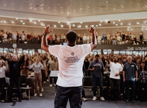Rear view of a speaker with raised hands facing a large standing congregation in a church auditorium