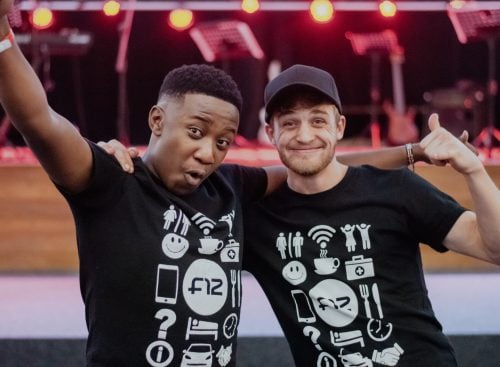 Two young men wearing matching black t-shirts with icons, smiling and posing with arms around each other in front of a stage
