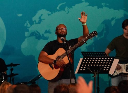 Male worship leader with acoustic guitar sings into a microphone, hand raised, band behind and audience in foreground