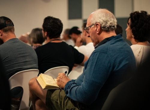 Older man in a blue jumper writing in a notebook while seated among other attendees