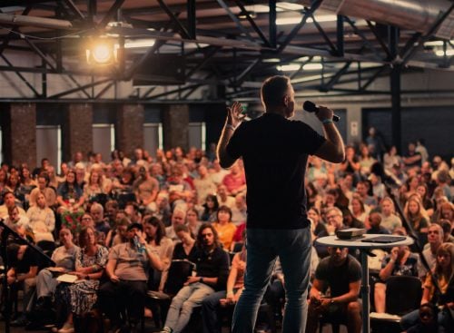 Speaker on stage addressing a large seated audience in an industrial-style hall