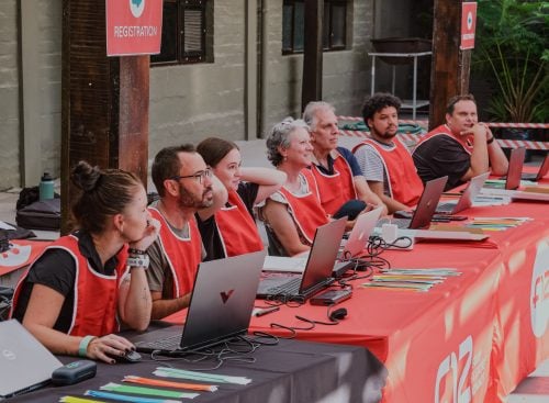 Row of registration volunteers in red vests seated behind a table with laptops and a 'REGISTRATION' sign