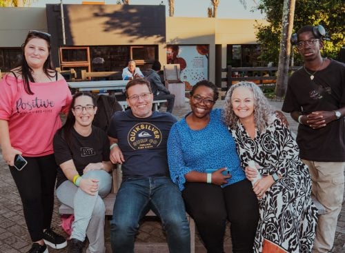 Six people smiling and posing outdoors on benches and standing in a courtyard
