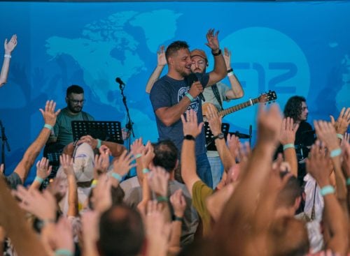 Speaker with microphone and band on stage leading a crowd with raised hands at an indoor event