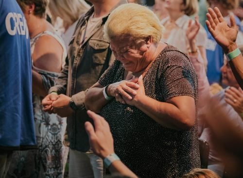 Elderly woman in a crowded congregation clasping her hands to her chest with eyes closed