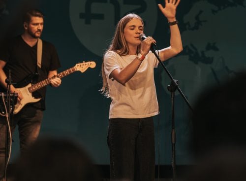 Female singer on stage holding a microphone and raising one hand, with a guitarist playing behind her and audience in