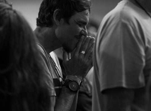 Man in a crowd with hands clasped near his mouth and eyes closed, appearing in quiet reflection