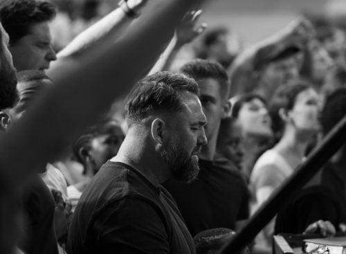 Black and white image of a crowd with raised hands; a bearded man stands with eyes closed and hands clasped