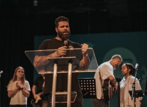 Bearded man speaker holding a microphone behind a clear lectern on stage with a worship band behind him