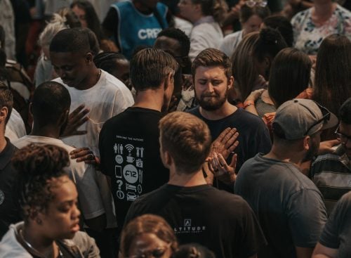 Crowded indoor gathering of people praying, many with hands on others' shoulders and eyes closed