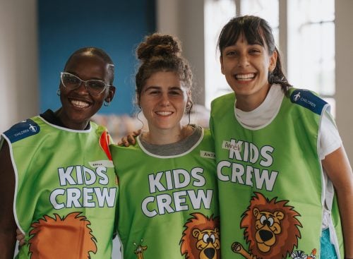 Three smiling adults wearing green 'Kids Crew' vests with lion graphic, standing together indoors