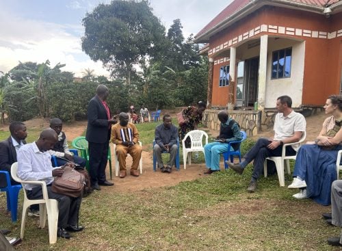 Group of people seated in plastic chairs outdoors beside a building, one person standing and speaking