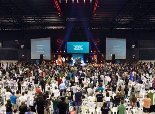 Large indoor gathering with a person on stage leading a crowd standing before three screens and stage lights