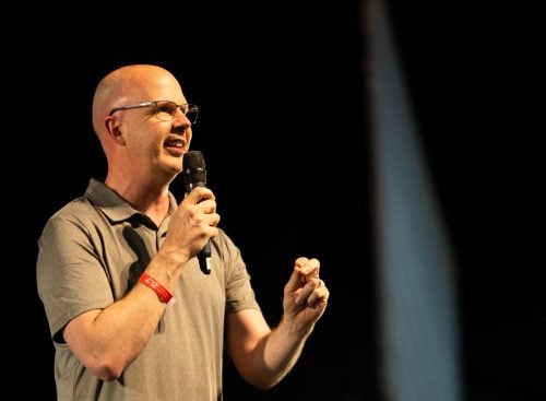 Bald man wearing glasses and a grey polo speaking into a microphone on a dark stage, red wristband visible