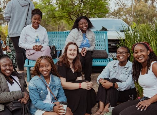 Group of eight young women sitting and kneeling by a blue bench outdoors, smiling at the camera